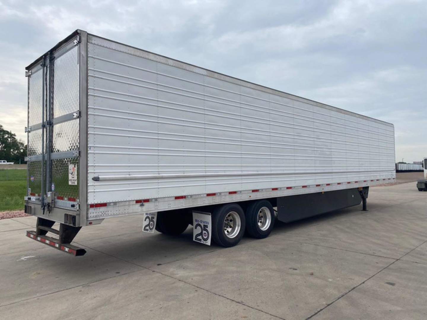 Utility reefer trailer showing triple-axle wheels, aluminum side panels, rear ramp doors, and a small label near the left rear corner.
