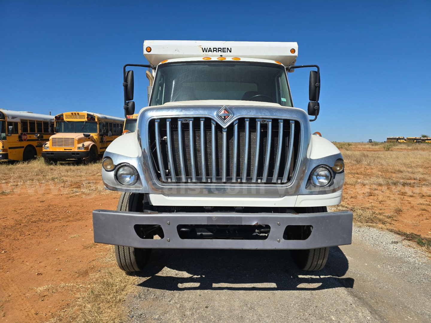 Front view of a 2013 International 7400 dump truck with large chrome grille and bumper, Warren header, in a dirt lot.