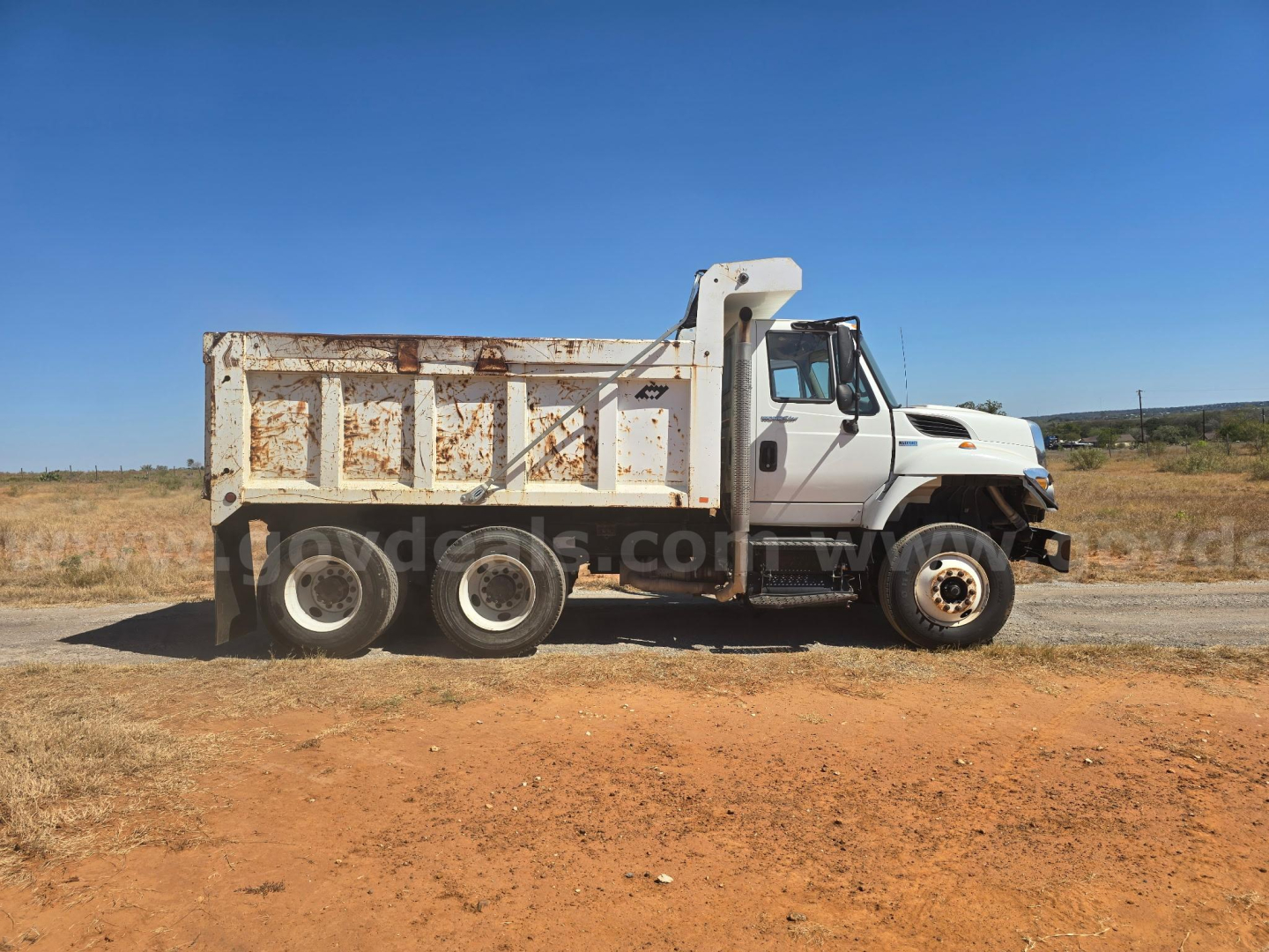 Dump truck side view showing worn body with rust highlights and three-axle rear. International 7400 dump truck.