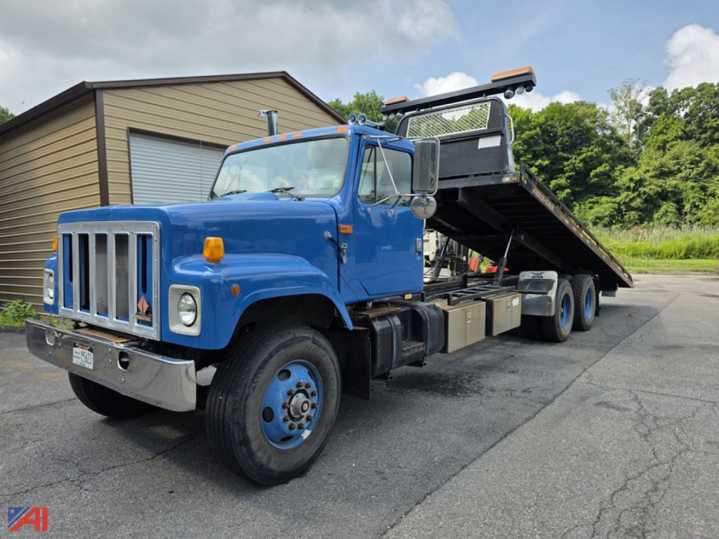 Blue 2000 International 2574 rollback truck with a tilting flatbed and elevated winch platform, front grille and bumper visible, white cab and blue body, on asphalt.