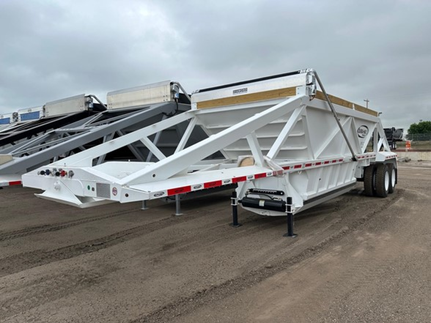 Bottom dump trailer in white with truss frame showing sturdy steel structure and dual rear axles; exterior side rails and loading chute visible.