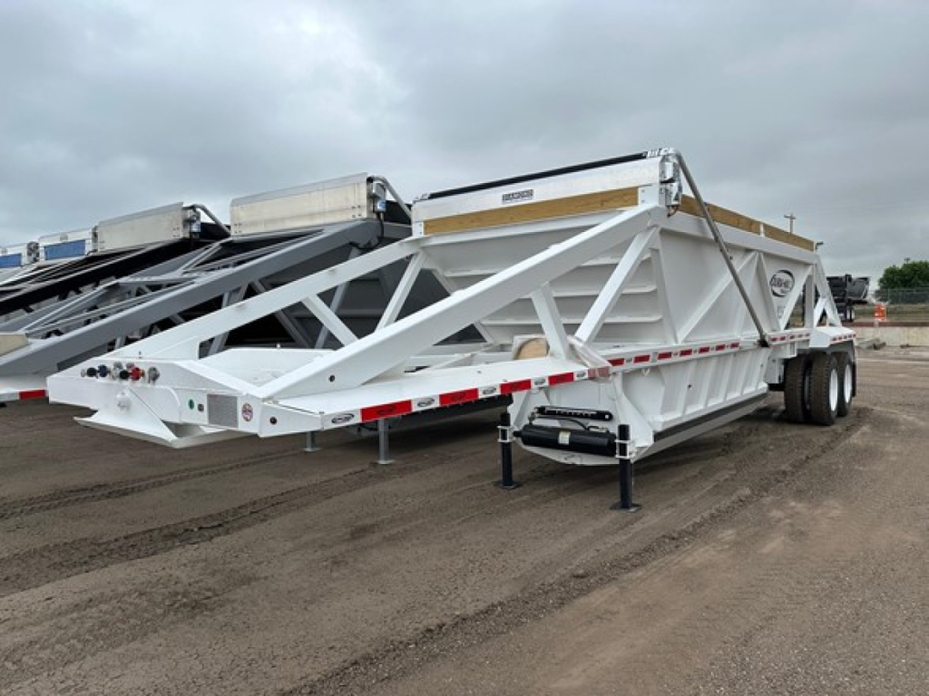 Trailer side view showing a white Dura Haul bottom-dump trailer with framed lattice structure and dual rear axles, label panels, and support legs.