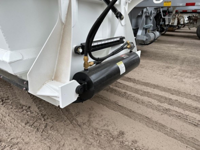 Bottom dump trailer corner showing hydraulic cylinder and tubing with black hydraulic reservoir and white metal frame.