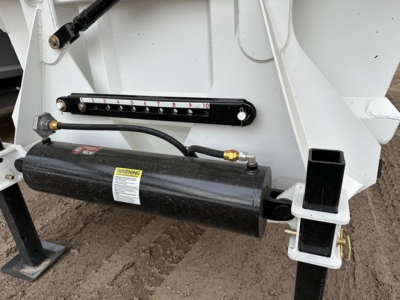 Trailer bottom dump hinge area showing hydraulic cylinder with warning label, level indicator gauge, and frame mount.