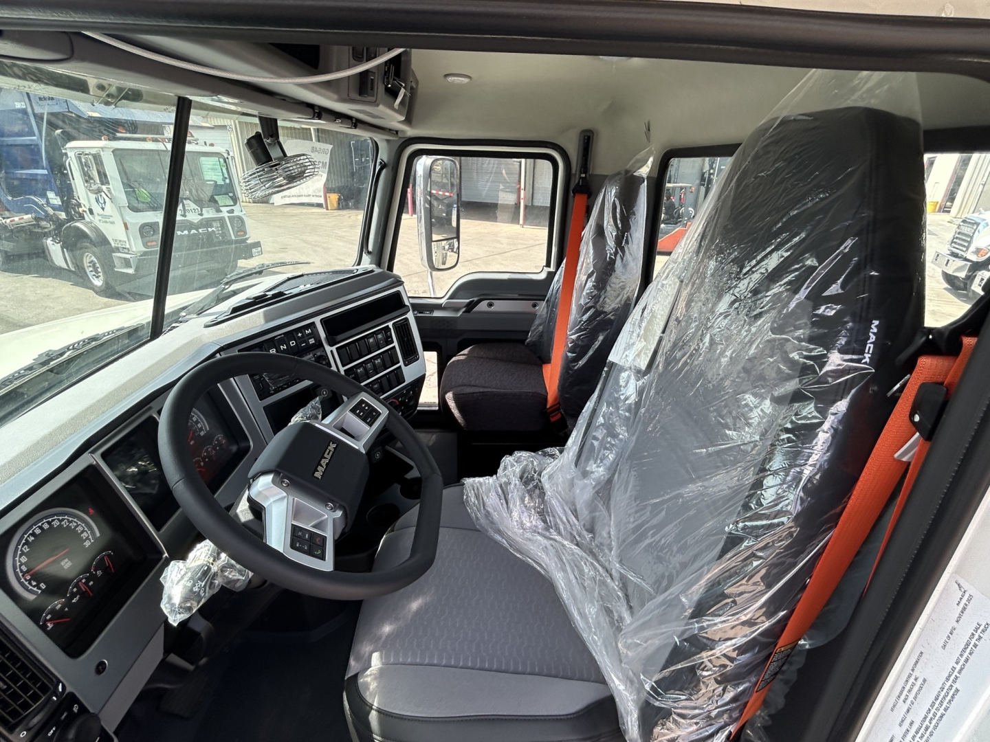 Mack GR64B cab interior showing steering wheel, dashboard, driver seat and passenger seat wrapped in plastic, with gray upholstery and orange seat belts.