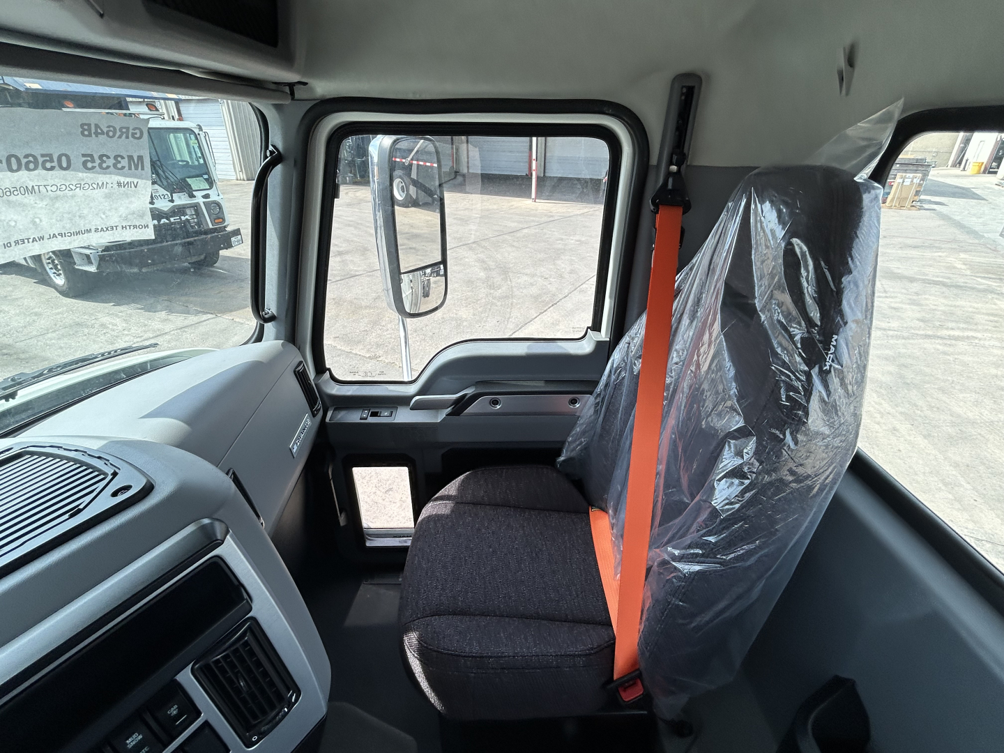 Cab interior of a new 2026 Mack GR64B rolloff truck showing gray seat with orange seatbelt, dashboard, and windshield view.
