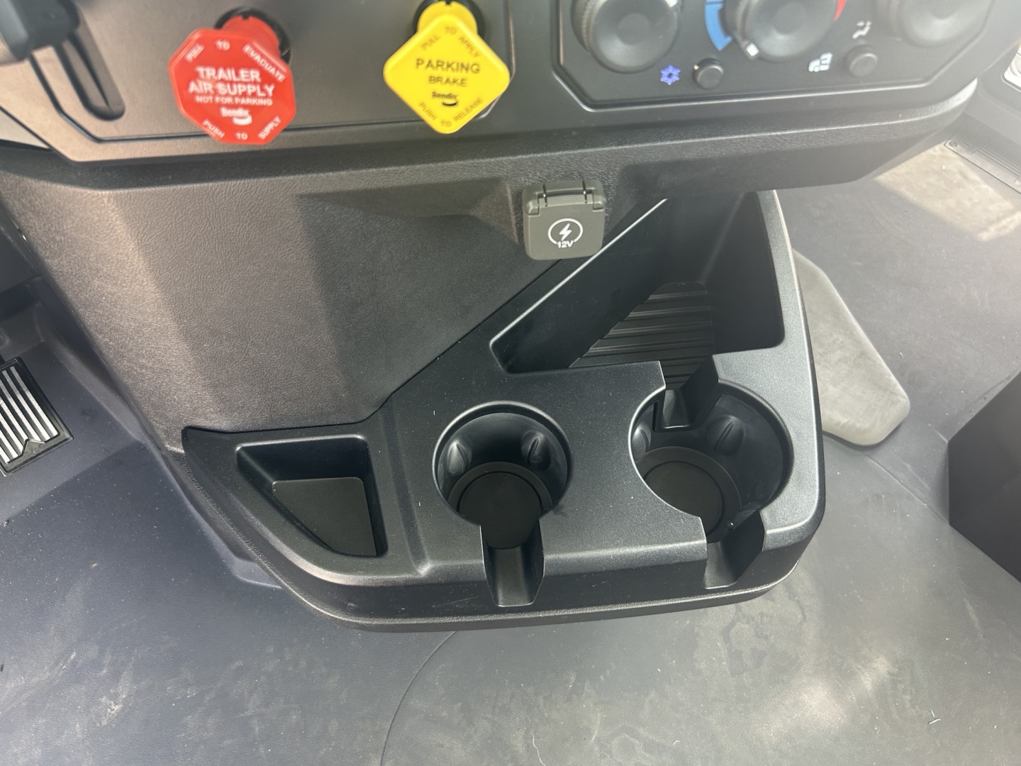 360-degree view of Mack GR64B dash area showing two cup holders, a 12V power outlet, ignition/controls, and the lower center console with a hazard switch and passenger footwell.
