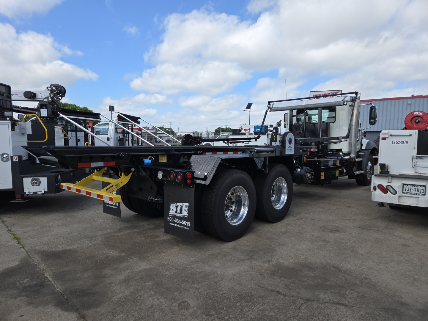 Mack GR64B roll-off truck rear view showing dual rear axles with heavy-duty tires and equipment platform; visible text on mudflap and rear bumper: BTB? 71678 dealership markings; clear blue sky with other trucks in background.