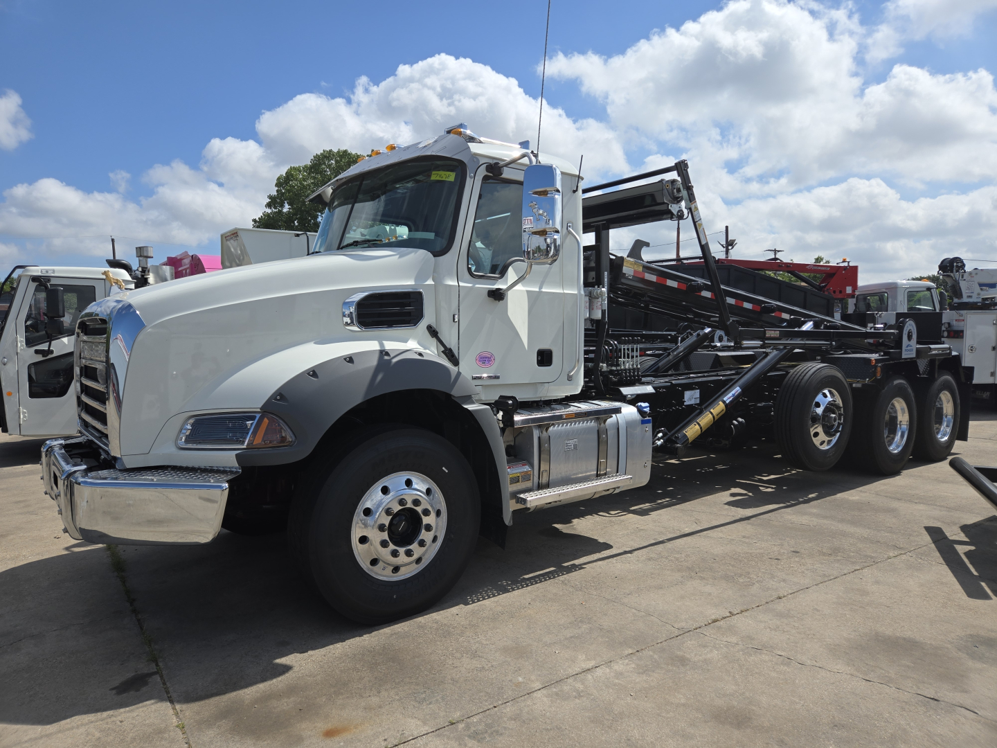 Mack GR64B rollover truck in a dealership lot showing front left side and hydraulic arm on flatbed chassis with chrome steps and black tires.