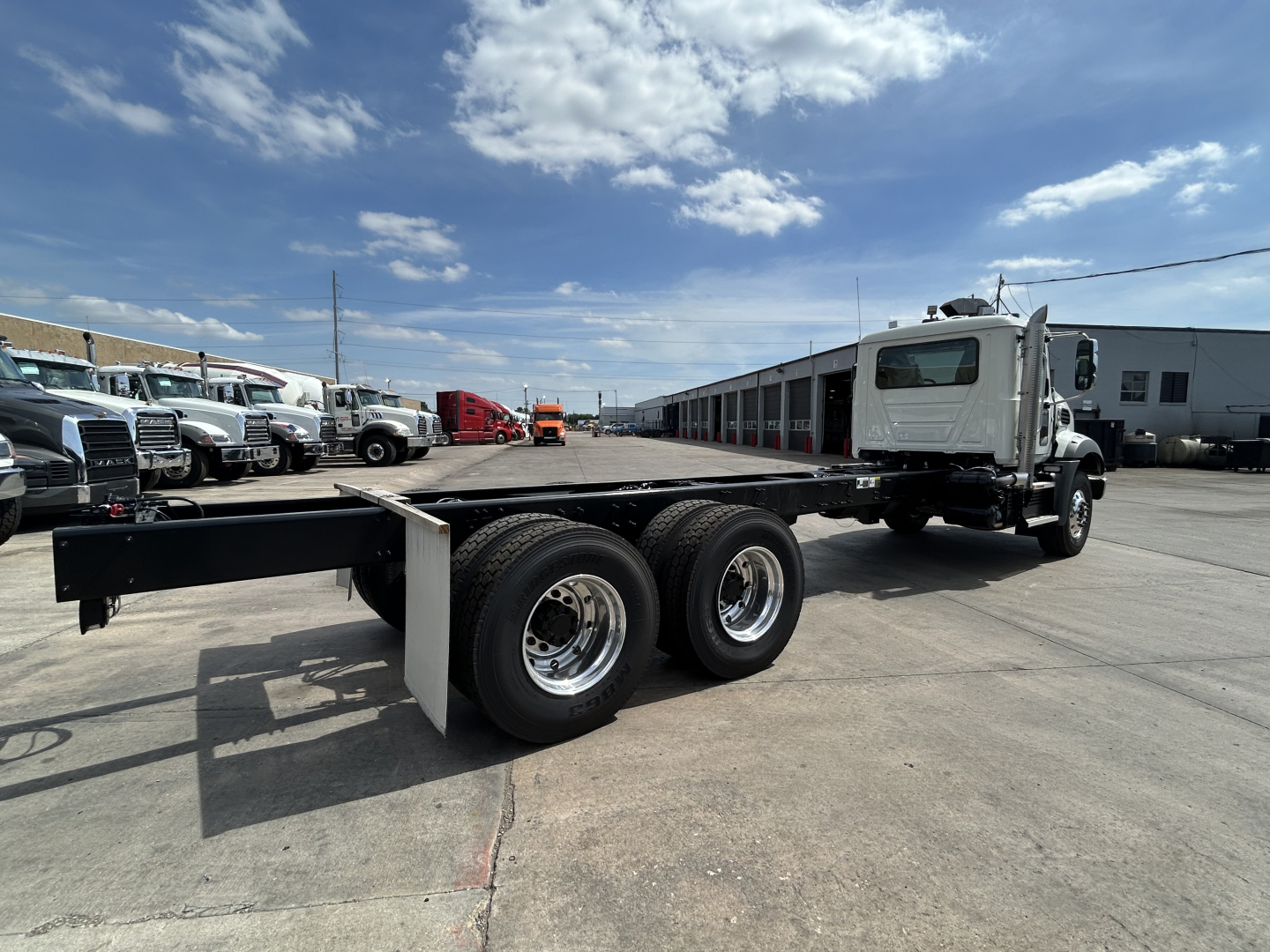 Truck chassis of a new Mack GR64B rolloff truck showing dual rear axles and flatbed frame with white cab in background.