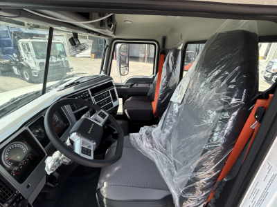 Mack GR64B cab interior showing steering wheel, dashboard, driver seat and passenger seat wrapped in plastic, with gray upholstery and orange seat belts.