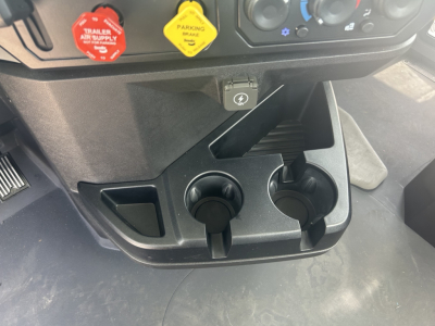 360-degree view of Mack GR64B dash area showing two cup holders, a 12V power outlet, ignition/controls, and the lower center console with a hazard switch and passenger footwell.