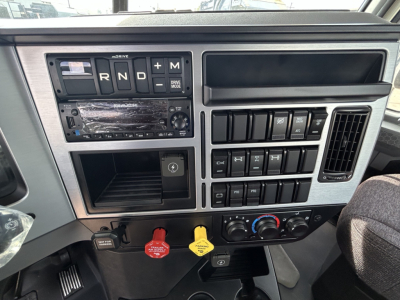 Truck dashboard of a 2026 Mack GR64B showing gear selector R N D M, multimedia head unit, climate controls, and warning/auxiliary switches.