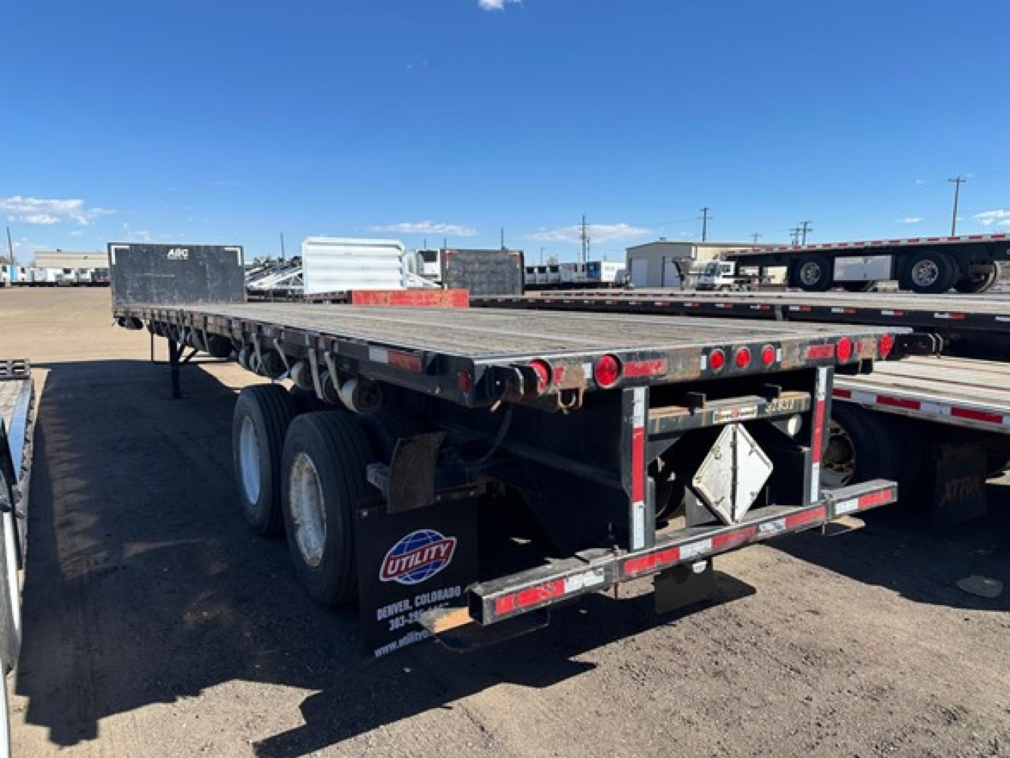 Trailer rear view showing flatbed with dual rear axles and mud flaps, red tail lights, and reflective tape along edges.