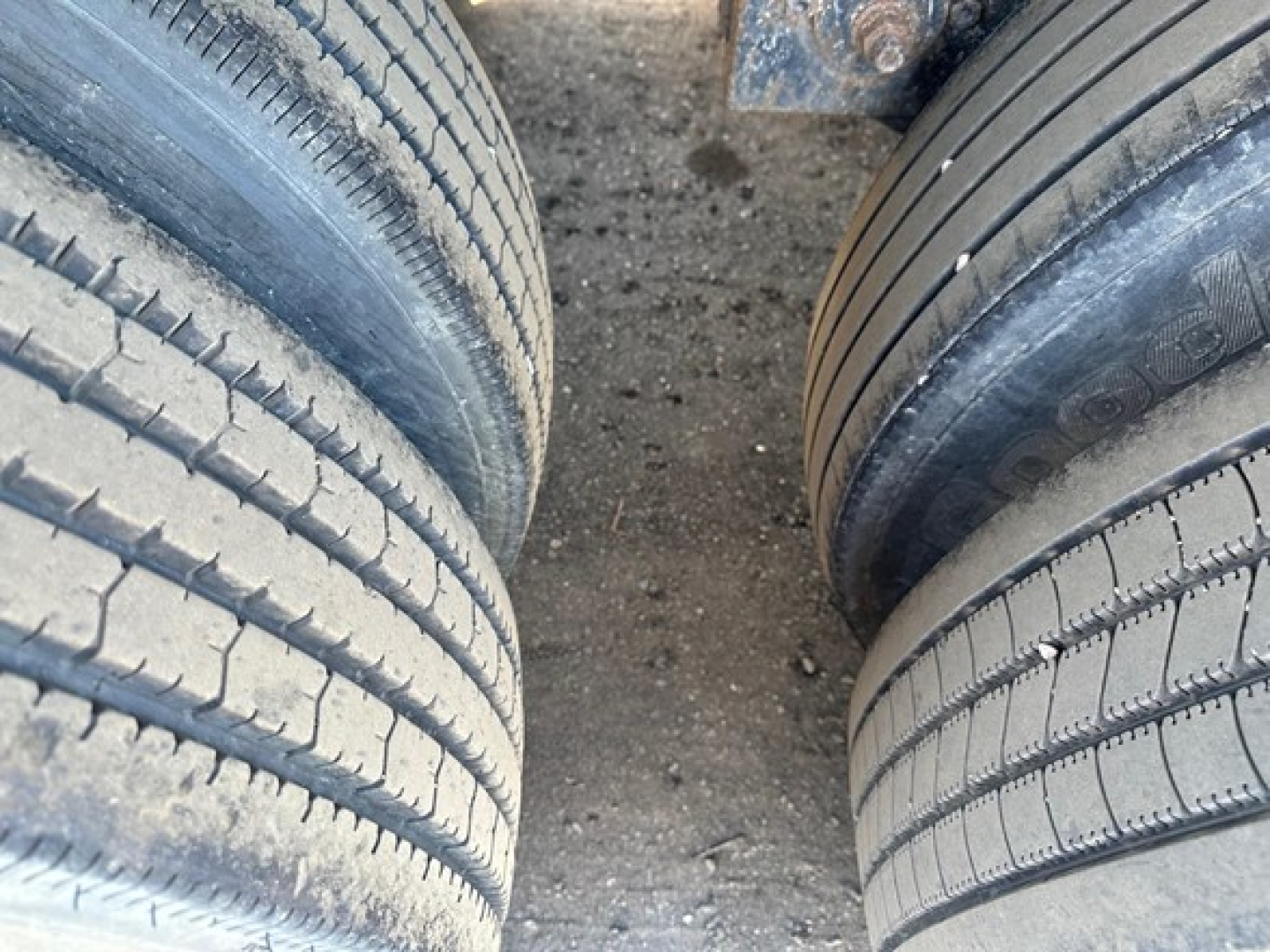 Flatbed trailer axles with dual tires showing tread and dirt surface.