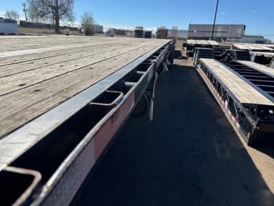 Flatbed trailer deck with metal frame and wheel wells, long wooden planks, and multiple trailers parked in lot.
