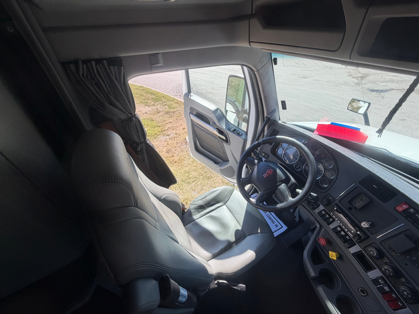 Cab interior of a Kenworth T680 truck showing driver’s seat, steering wheel with dashboard gauges, and open door; windows reveal outside scene.