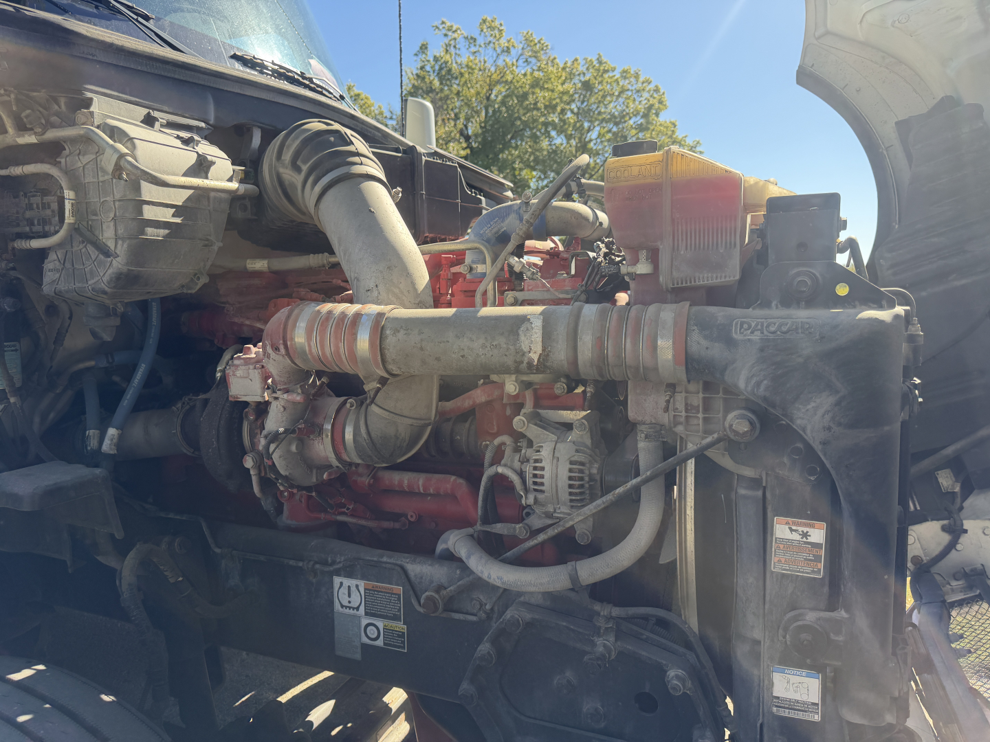 Kenworth T680 engine bay showing coolant reservoir, hoses, turbo piping, alternator, and radiator area with various stickers and labels.