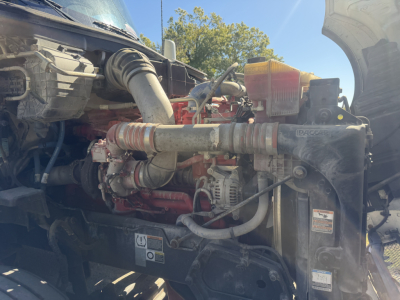 Kenworth T680 engine bay showing coolant reservoir, hoses, turbo piping, alternator, and radiator area with various stickers and labels.