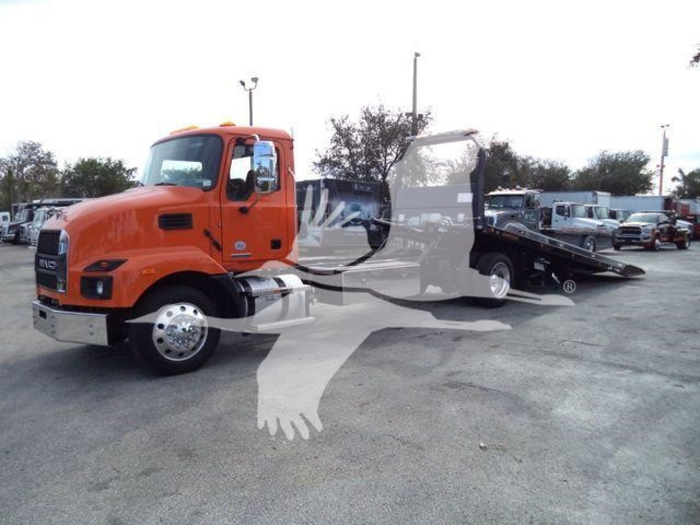 Orange 2026 Mack MD6 rollback tow truck in a dealership lot with a deployed flatbed and other trucks in background.