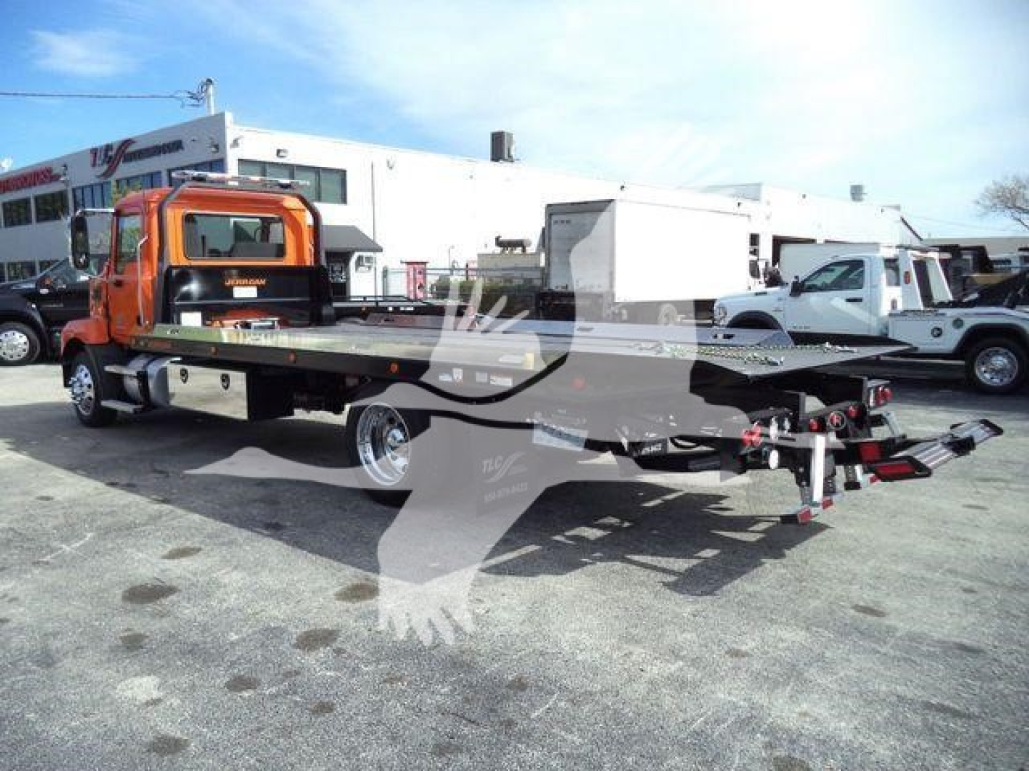 Flatbed tow truck chassis with orange cab showing hydraulic deck tilt and winch in a dealership lot.