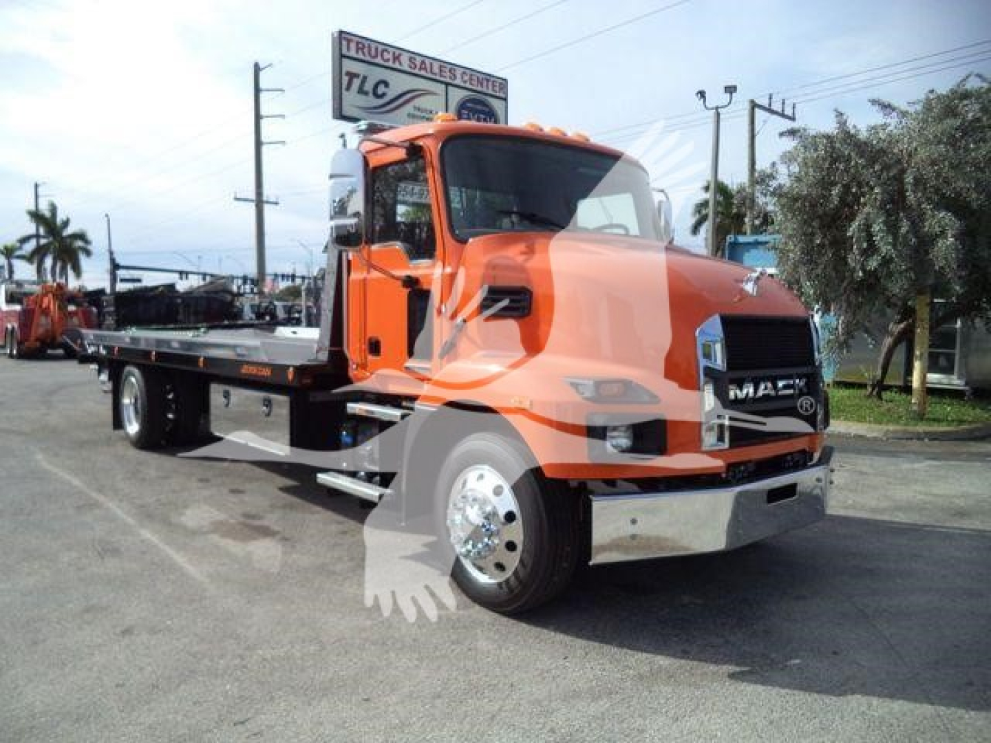Mack MD6 tow truck in orange on a dealership lot, front grille with Mack emblem, long flatbed, TLC Truck Sales Center sign in background.