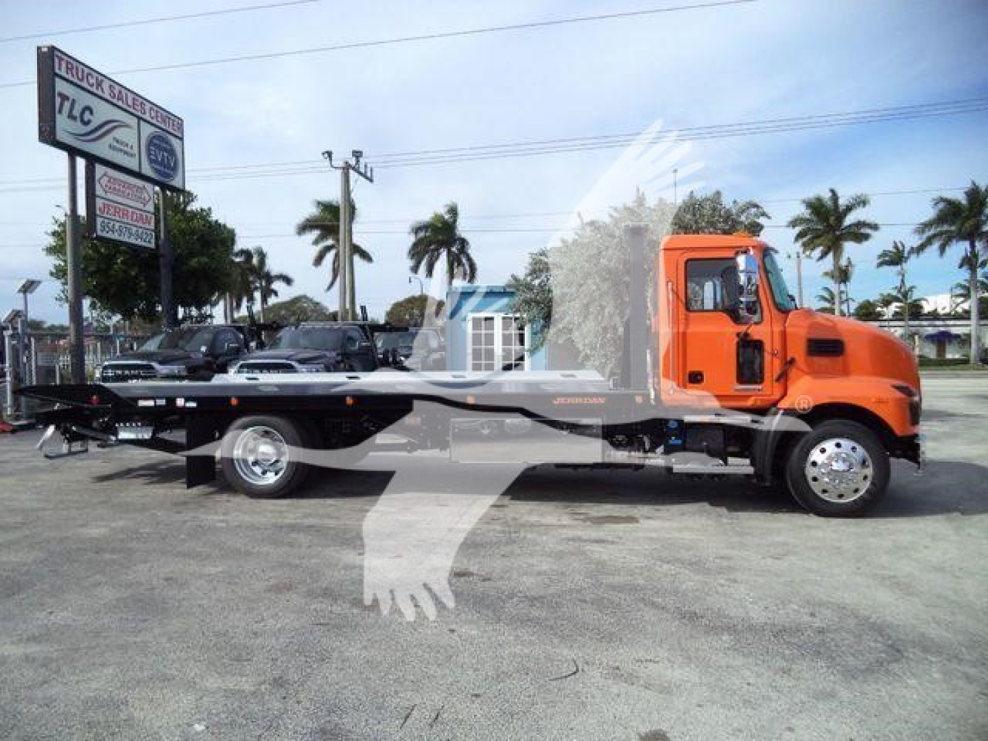MD6 rollback tow truck in orange cab with flatbed, side view showing trailer hitch and wheels, dealership lot in background