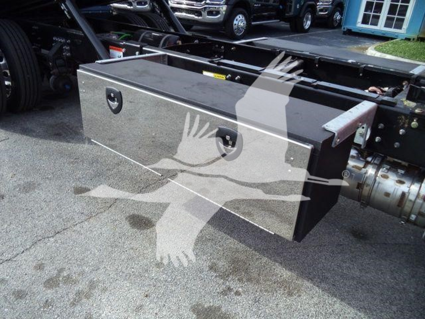 Large aluminum toolbox mounted under Mack MD6 truck frame with two latches, reflective surface, and mounting brackets visible.