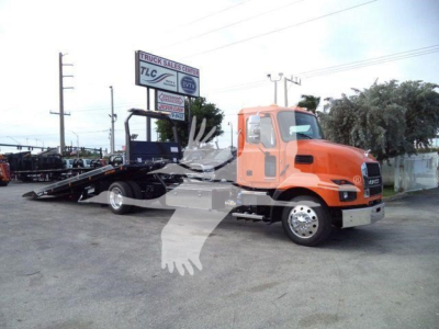 2026 Mack MD6 roll-back tow truck in orange cab with flatbed lifted for loading, side view showing wheel tips and truck dealership in background.