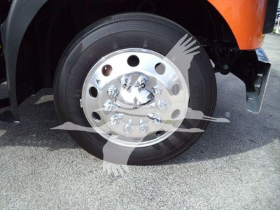Wheel end of a 2026 Mack MD6 truck showing a polished aluminum hub with multiple lug nuts and a black tire, part of an orange body panel visible.