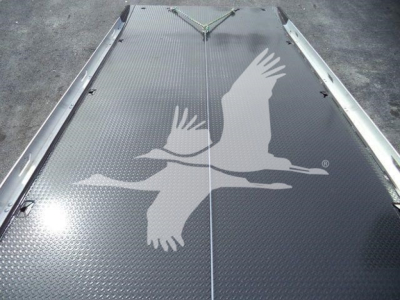 Diamond-plate metal platform with a white flying geese logo across the surface, inset lines and tie-down hooks at the edges; truck equipment deck image.