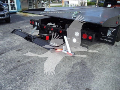 Flatbed tow truck rear view with extendable wheel-lift and ramps, black chassis, multiple red taillights, and chain lasso visible.