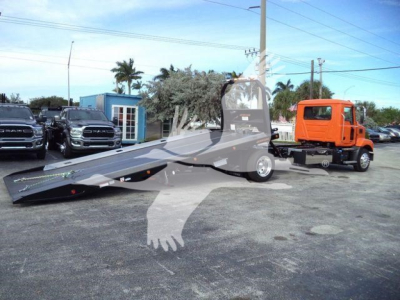 MD6 tow truck on trailer in lot with orange cab, roll-back flatbed extended, other trucks in background.