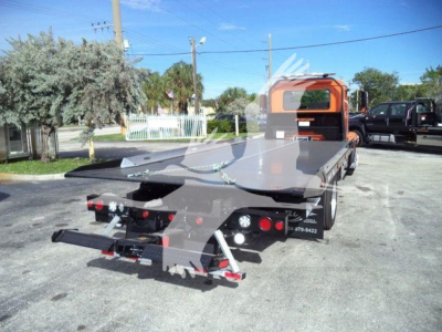 Truck bed trailer and cab of a Mack MD6 rollback tow truck with orange cab and grey flatbed, rear view showing tow apparatus and lights.