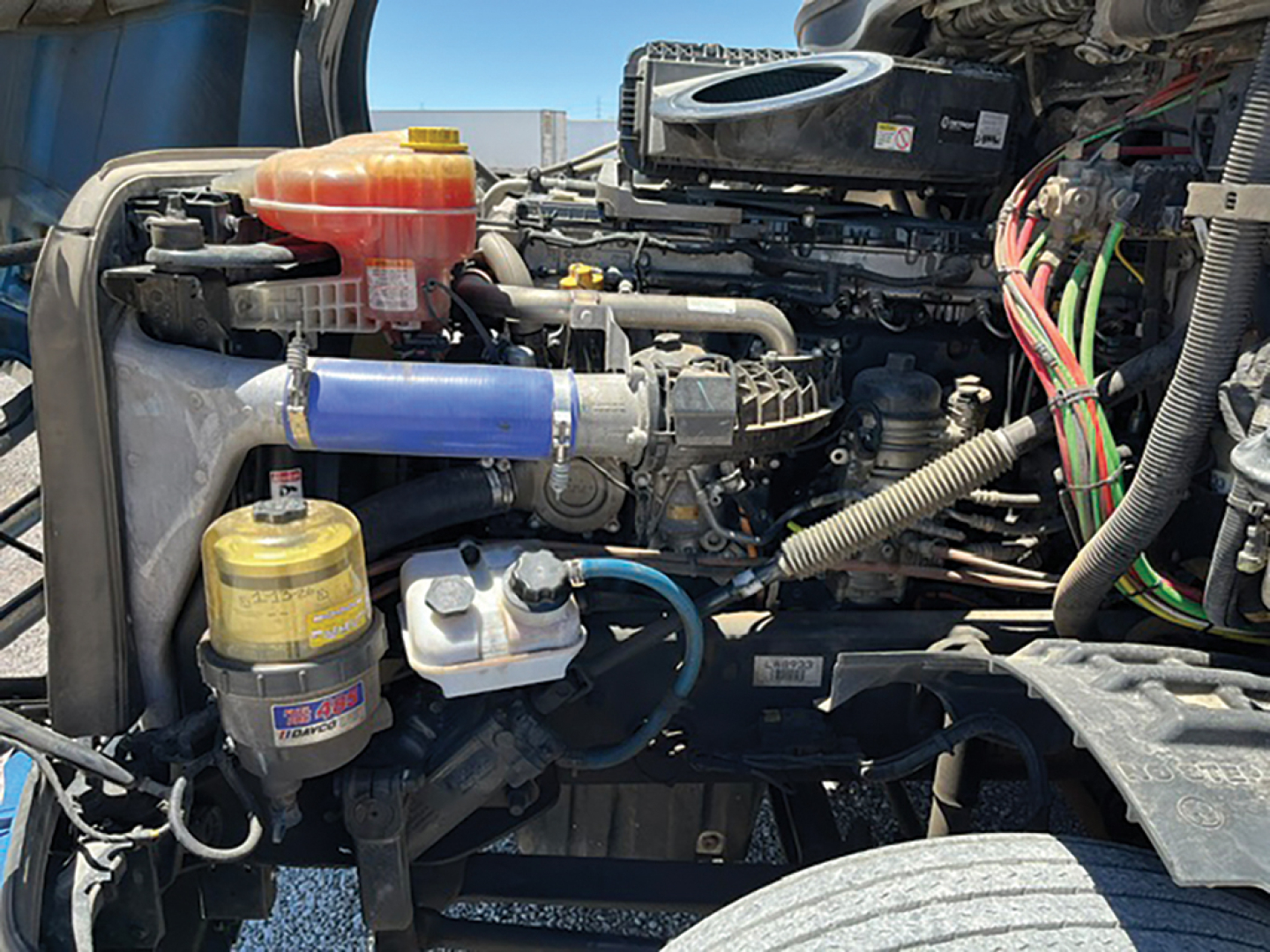Front section of a Freightliner Cascadia engine bay showing coolant reservoir, air filter canister, and blue intercooler hose with various wiring harnesses.