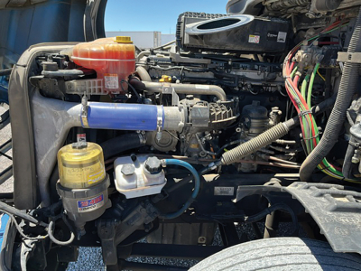 Front section of a Freightliner Cascadia engine bay showing coolant reservoir, air filter canister, and blue intercooler hose with various wiring harnesses.