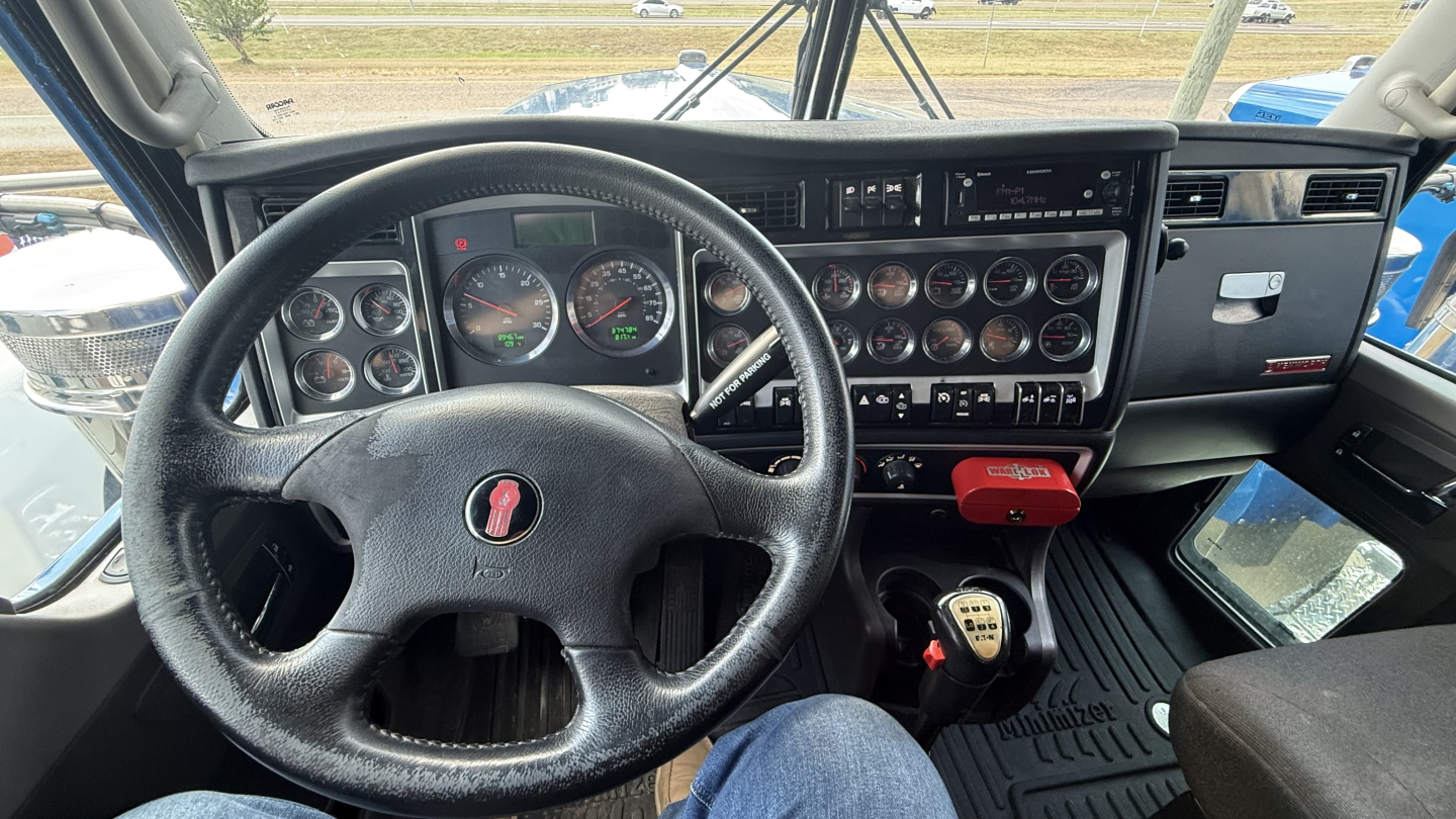 Kenworth W900 truck interior cockpit showing steering wheel, gauge cluster, and dashboard controls with gear shifter and switches.