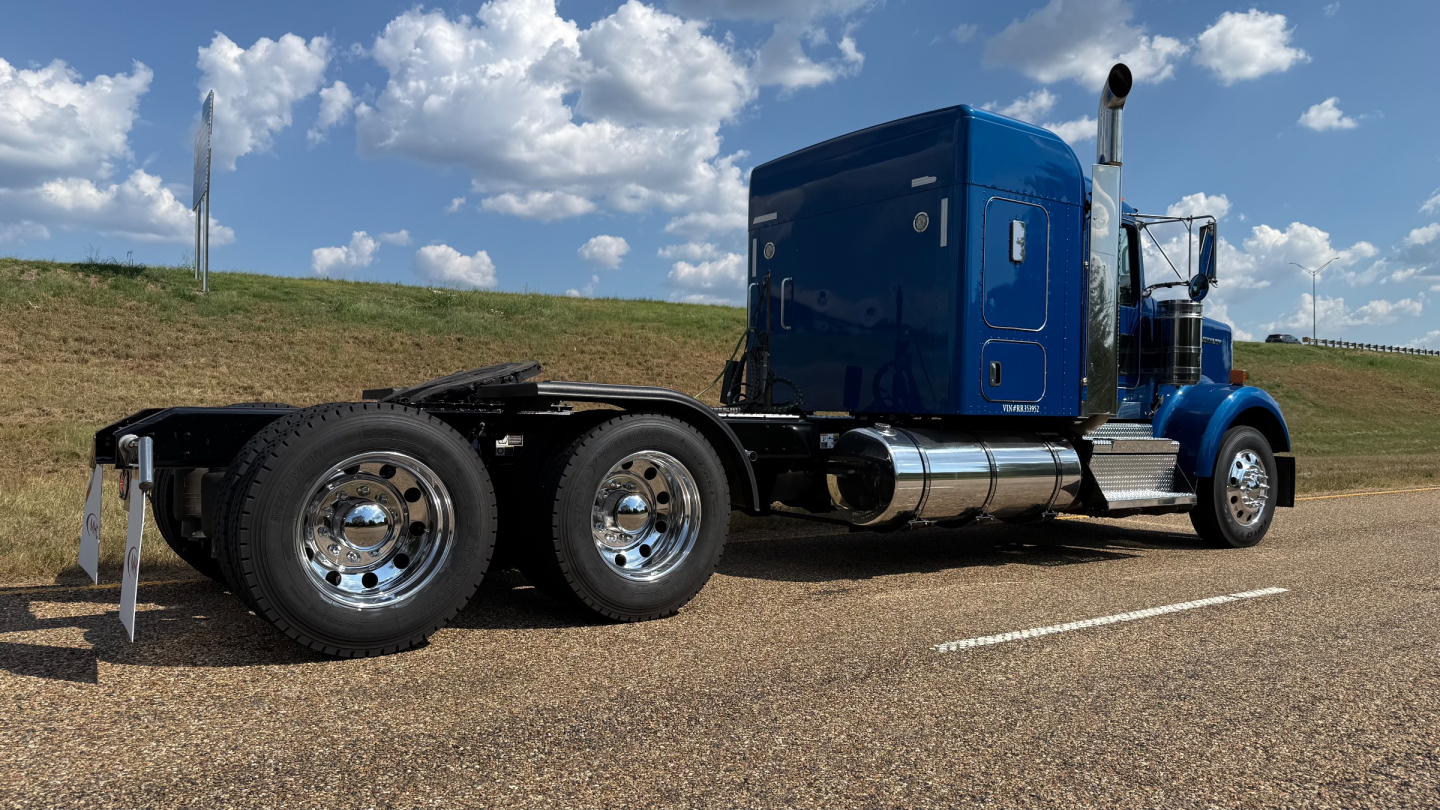 Kenworth W900 conventional sleeper tractor in blue, side view with dual rear axles and chrome fuel tanks.