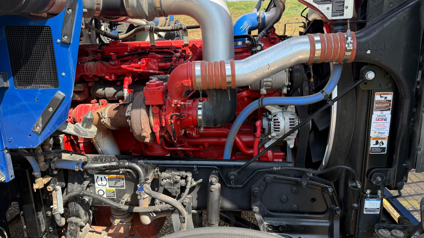 Kenworth W900 engine bay with red engine, silver turbo pipes, blue accents, and various hoses; visible components include hoses, belts, and an alternator.