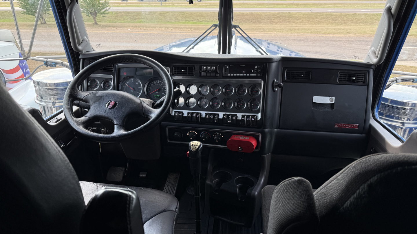 Cab interior of a Kenworth W900 truck showing steering wheel, dashboard with gauges, gear shifter, and center console.