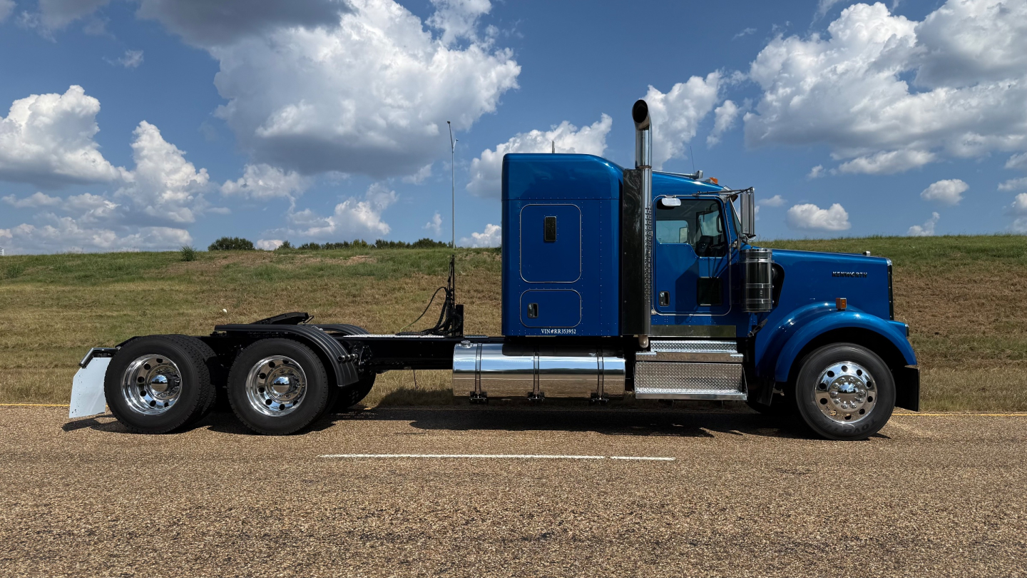 Kenworth W900 tractor side view showing blue cab, aluminum fuel tanks, and six-wheel rear chassis with chrome wheels.