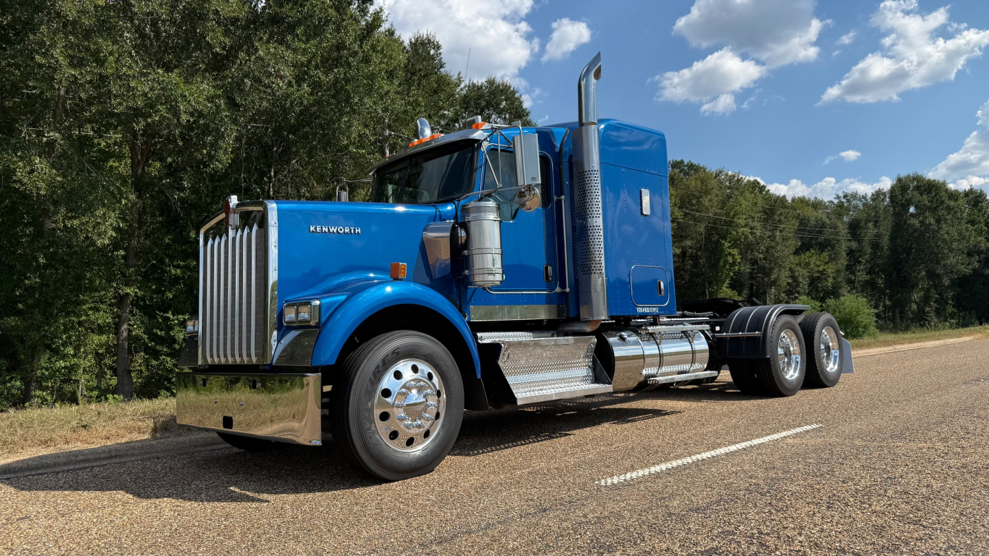 Kenworth W900 conventional sleeper truck in blue, side view, front grille and dual rear axles visible.