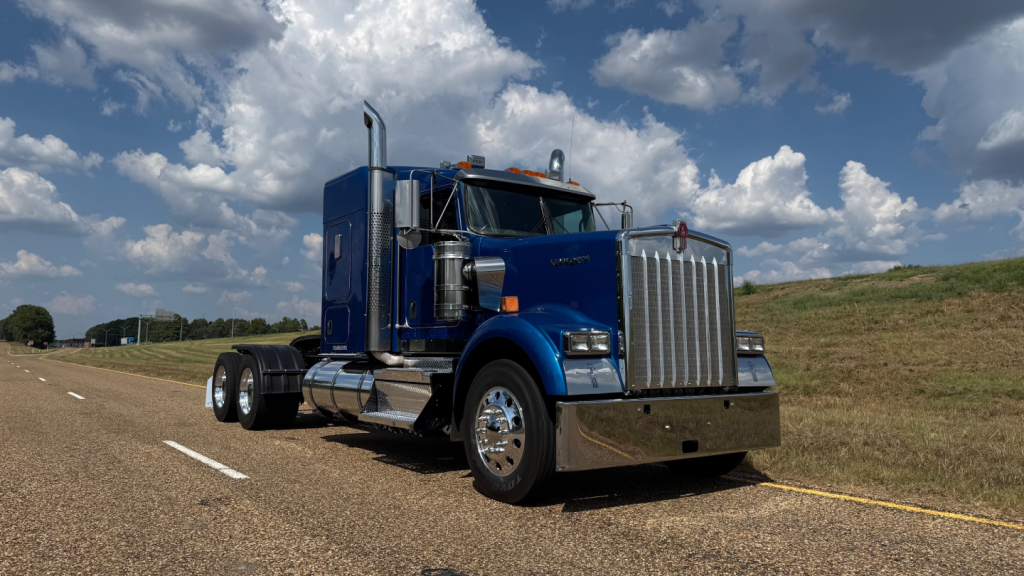 Kenworth W900 conventional sleeper truck in blue on highway showing chrome front grille and dual rear axles.