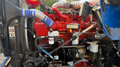 Kenworth W900 engine bay with red X15 by Cummins-style badge, multiple hoses, filters, and blue intake hose in view.