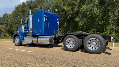Kenworth W900 truck chassis with blue sleeper cab and dual rear axles, side view showing metal fuel tanks and exhaust stacks.