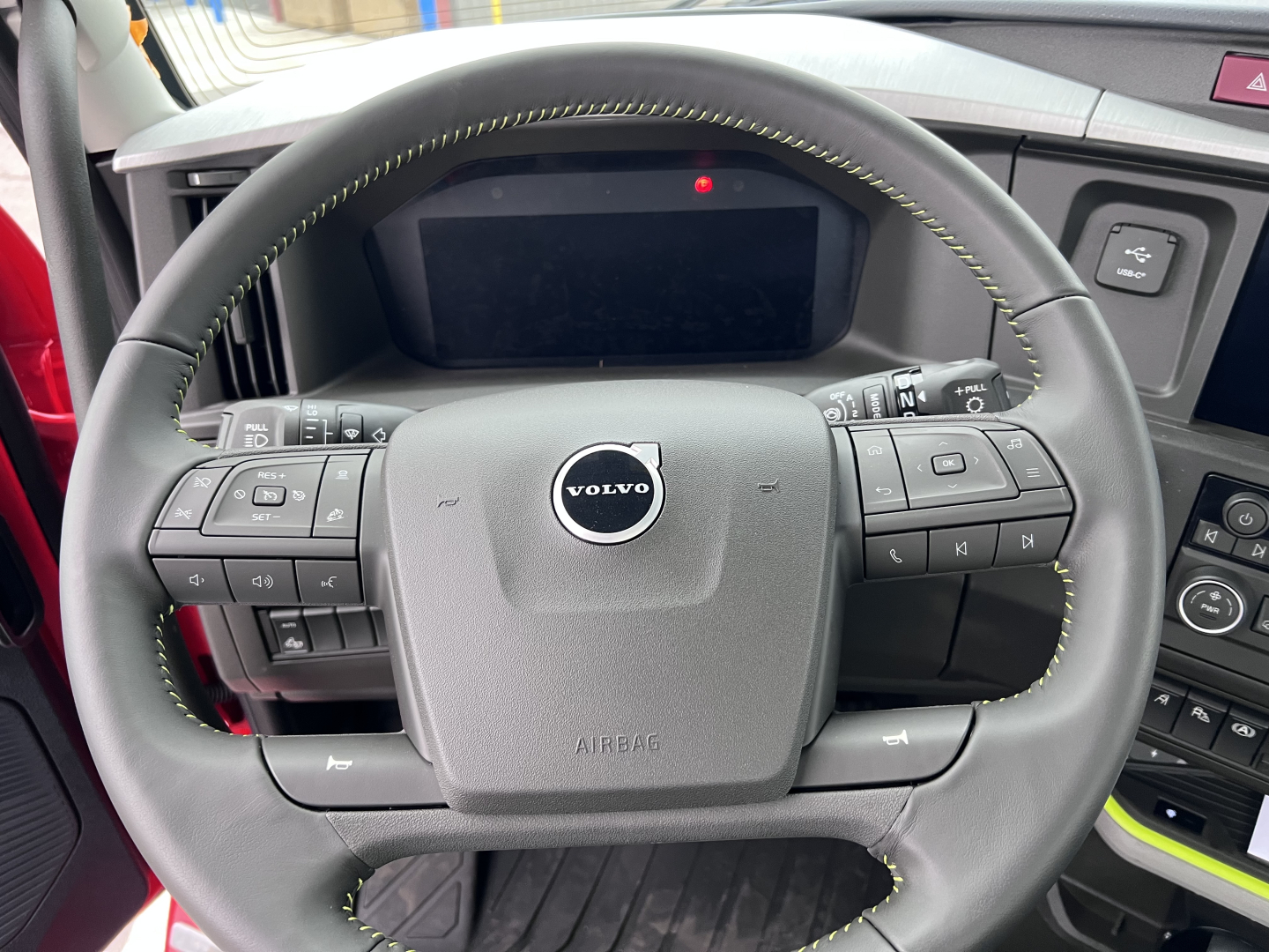 Volvo truck cabin steering wheel with dashboard and controls, Volvo logo on airbag, green stitching visible on steering wheel, digital display screen behind wheel.