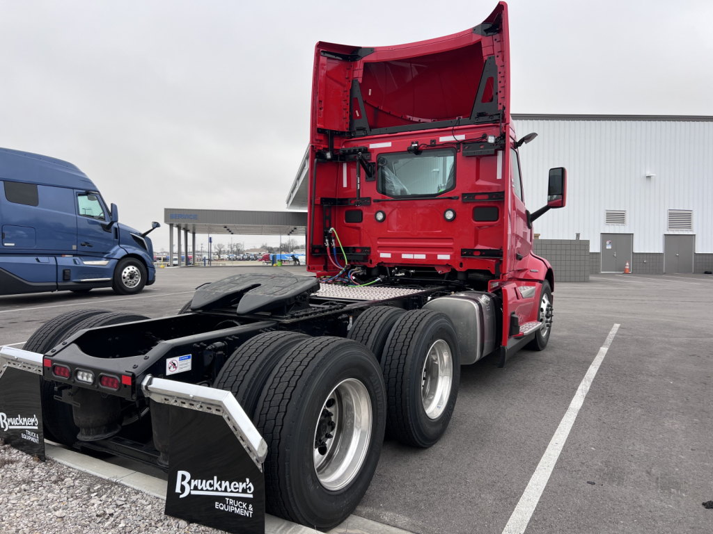 Truck tractor viewed from rear-left, red Volvlo VNL cab tilted with three rear axles, dual rear tires, and black fenders. Visible are metal plates, rear license area, and a white sticker near the mudflap.
