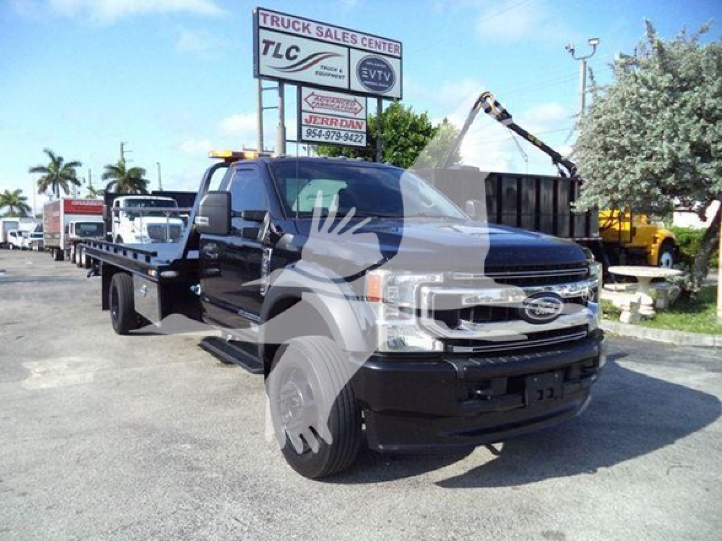 Ford F-600 tow truck in a lot with a flatbed and overhead light bar, front grille visible, dealership signage in background.