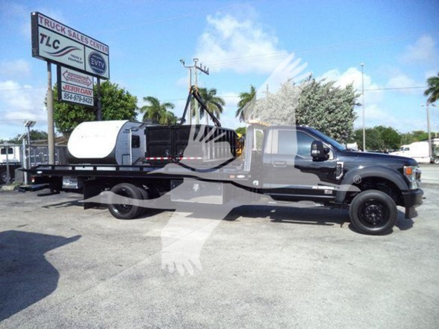 Ford F-600 rollback tow truck with a flatbed, black cab, and white mid-section, side view showing long bed and equipment; dealership lot in background.