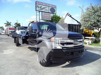 Ford F-600 tow truck in a lot with a flatbed and overhead light bar, front grille visible, dealership signage in background.